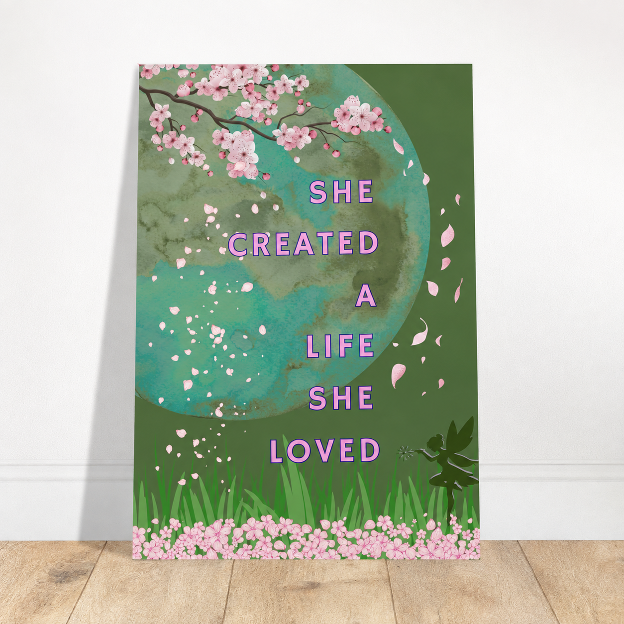 A vertical art print leaning against a white wall on a light wood floor. The print features a green background with a large teal-green circular graphic, pink cherry blossoms, green grass, and the text 'SHE CREATED A LIFE SHE LOVED' in pink.