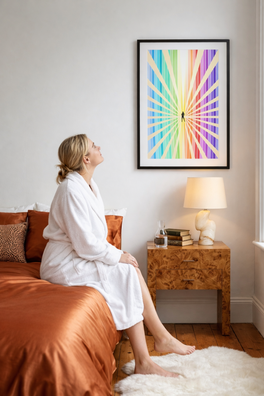 Framed Visibility feminist art print displayed above a bedside table in a soft neutral bedroom, with a woman sitting on the bed looking up at the artwork, styled for modern feminine interiors