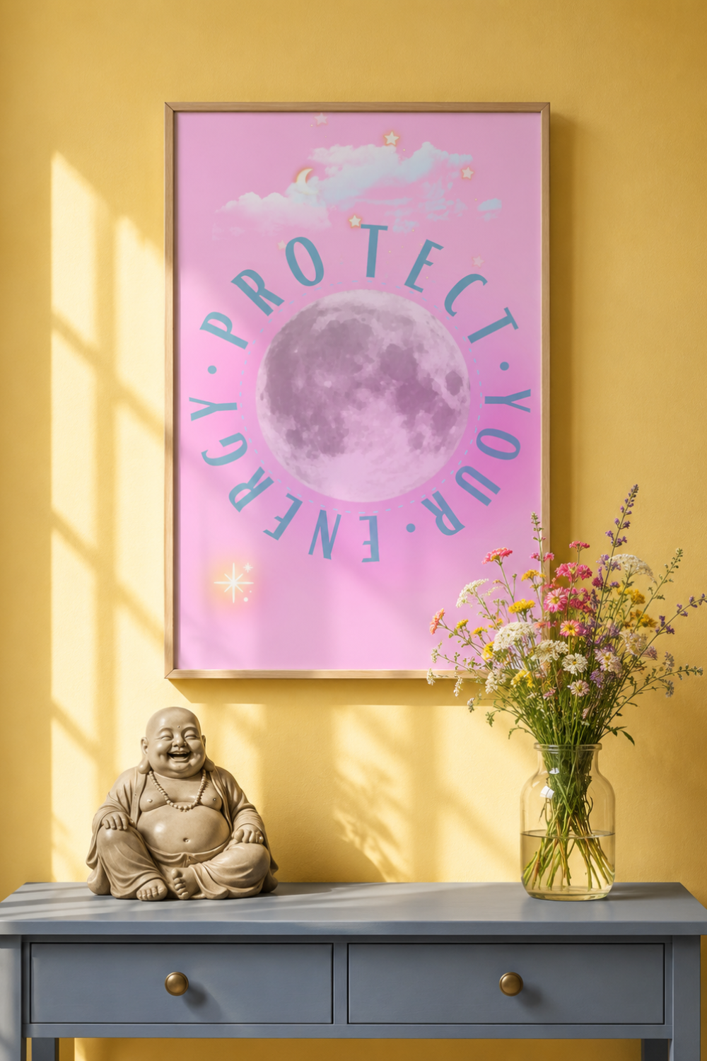 A framed art print featuring a pink background with a large moon graphic and the words 'PROTECT YOUR ENERGY' in blue, hanging on a yellow wall above a console table with a Buddha statue and a vase of flowers.
