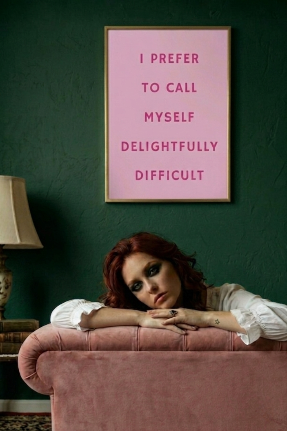 A framed pink typographic wall art print with the text 'I PREFER TO CALL MYSELF DELIGHTFULLY DIFFICULT' hangs on a dark green wall above a woman resting on a dusty pink sofa in a moody room.