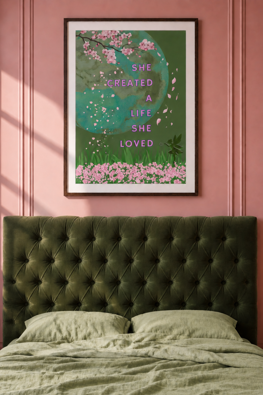 A framed art print featuring a green background, pink cherry blossoms, and the quote 'SHE CREATED A LIFE SHE LOVED' in pink text, displayed on a pink paneled wall above a dark green tufted bed headboard.