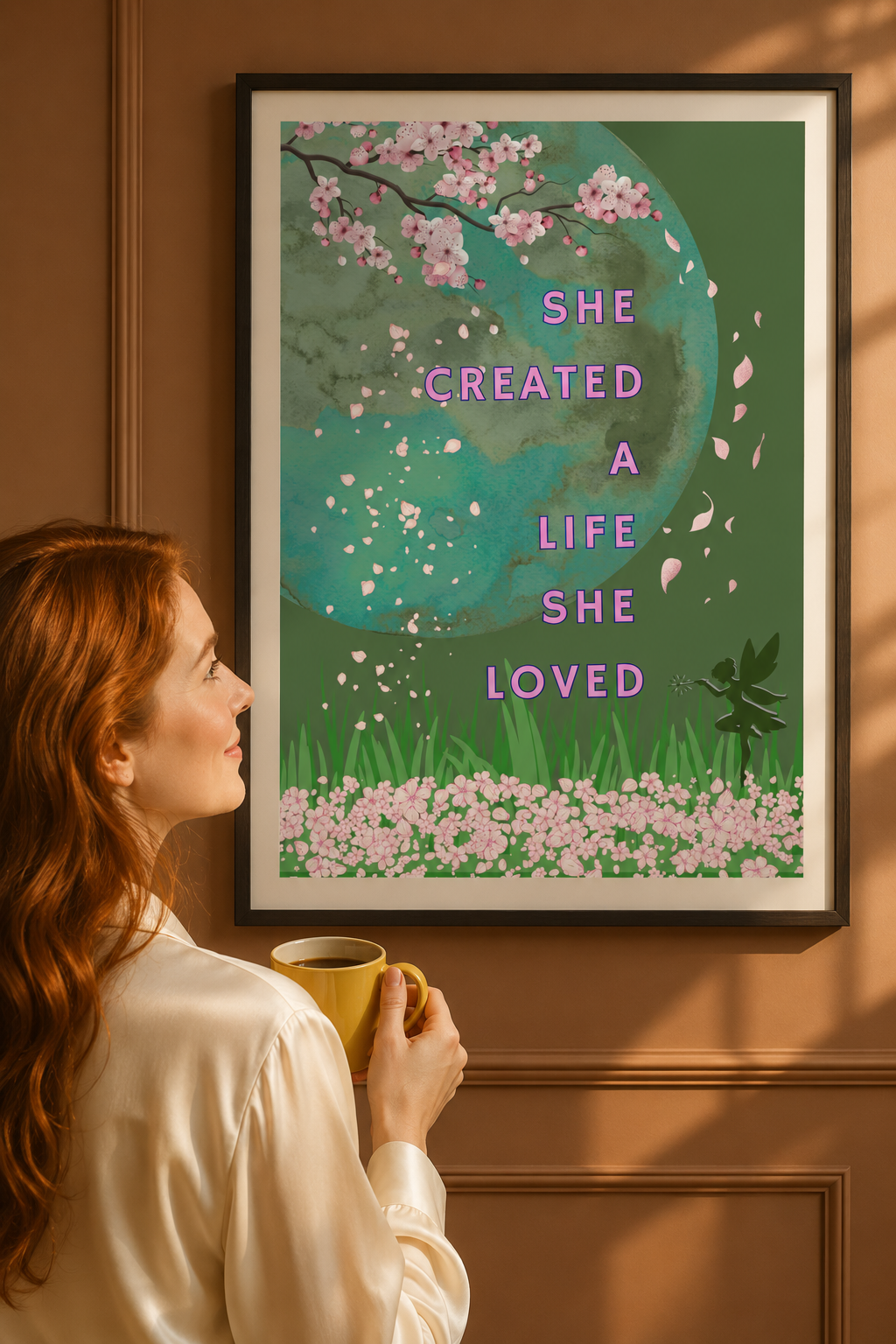 A framed art print featuring a green background with pink cherry blossoms, a teal moon, and the text 'SHE CREATED A LIFE SHE LOVED', hanging on a brown paneled wall. A woman with red hair in a white shirt holds a yellow mug, looking at the print.