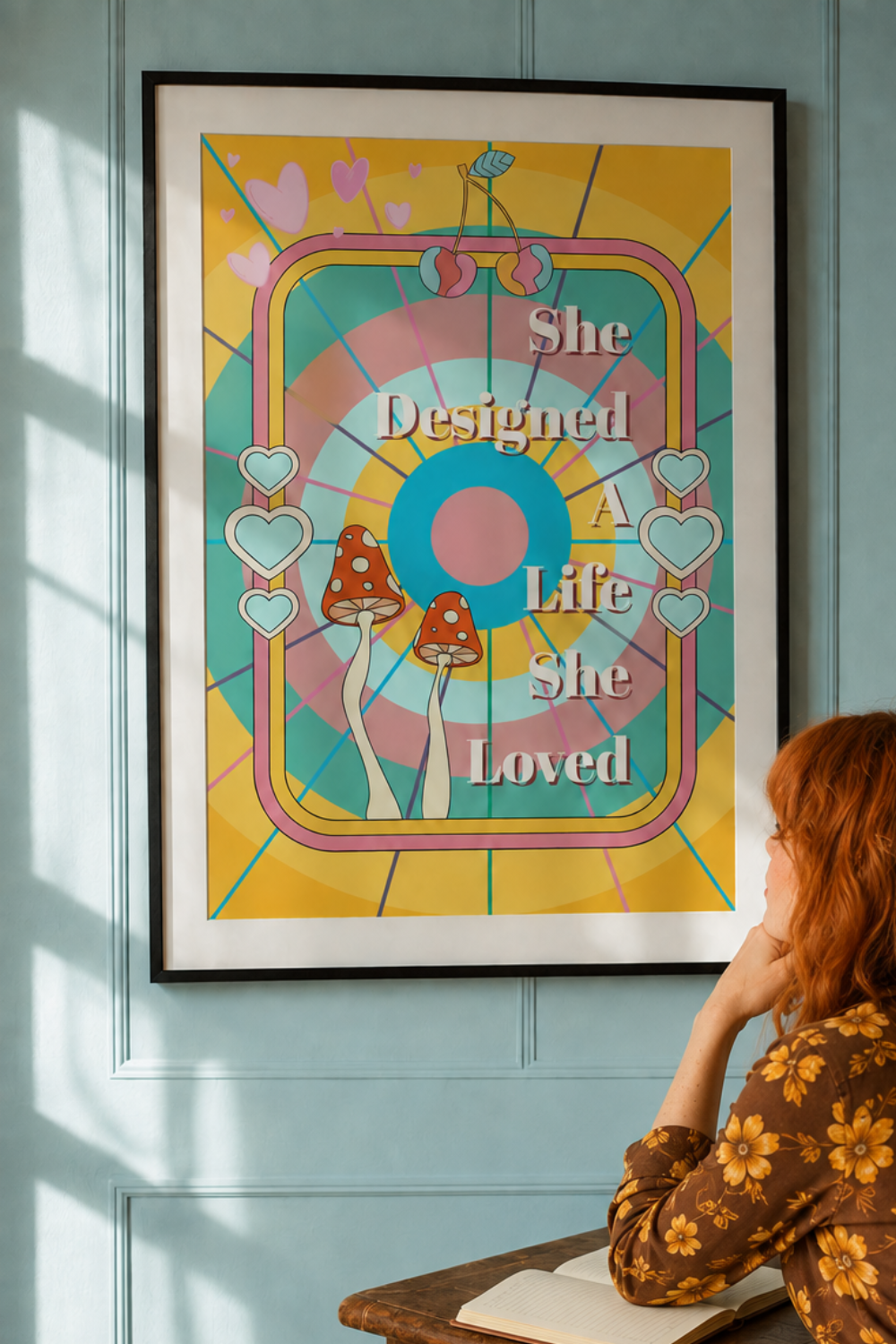 A framed art print with retro psychedelic graphics and the text 'She Designed A Life She Loved' is displayed on a light blue paneled wall. A woman with red hair sits at a wooden desk below, looking up at the print.