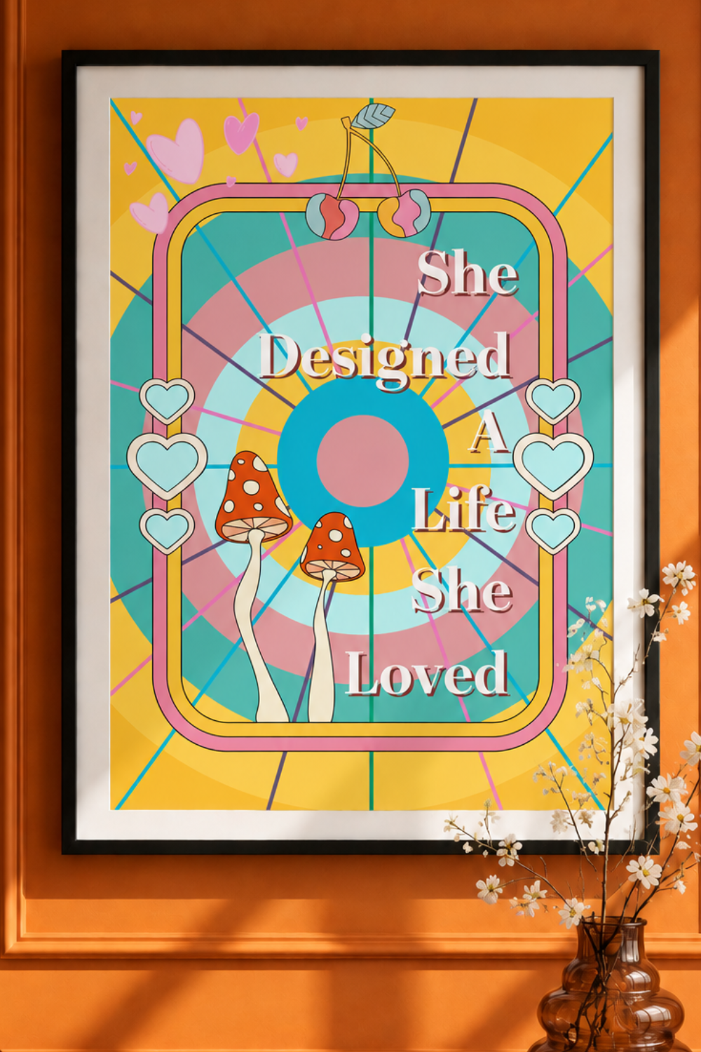 A framed art print with a colorful retro design featuring the text 'She Designed A Life She Loved' is displayed on an orange wall next to a vase with small white flowers.