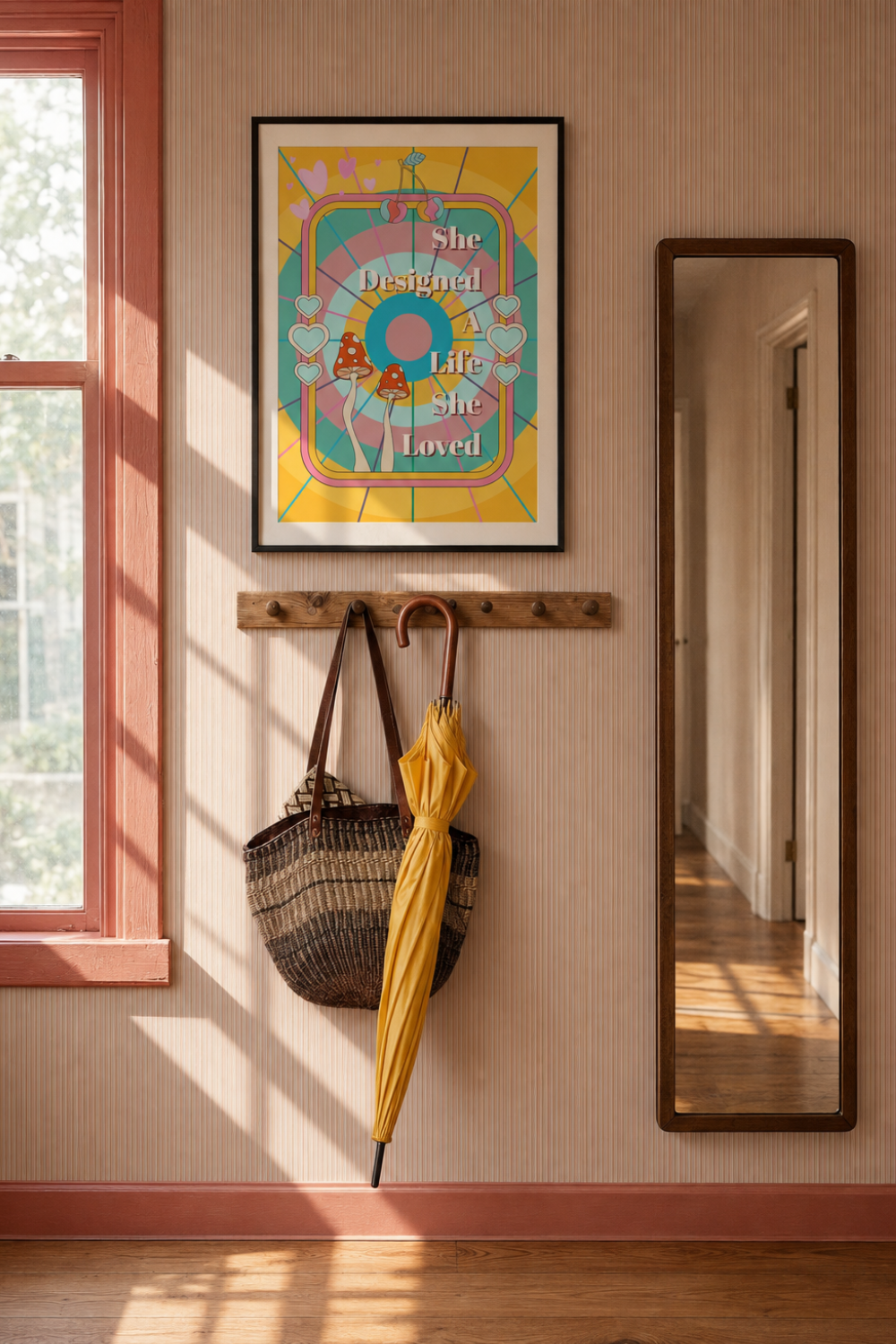 A framed art print with She designed a life she loved a colourful retro design hangs on a beige wall in a sunlit room. Below the print, a wooden coat rack holds a woven tote bag and a yellow umbrella, with a tall floor mirror reflecting a hallway.