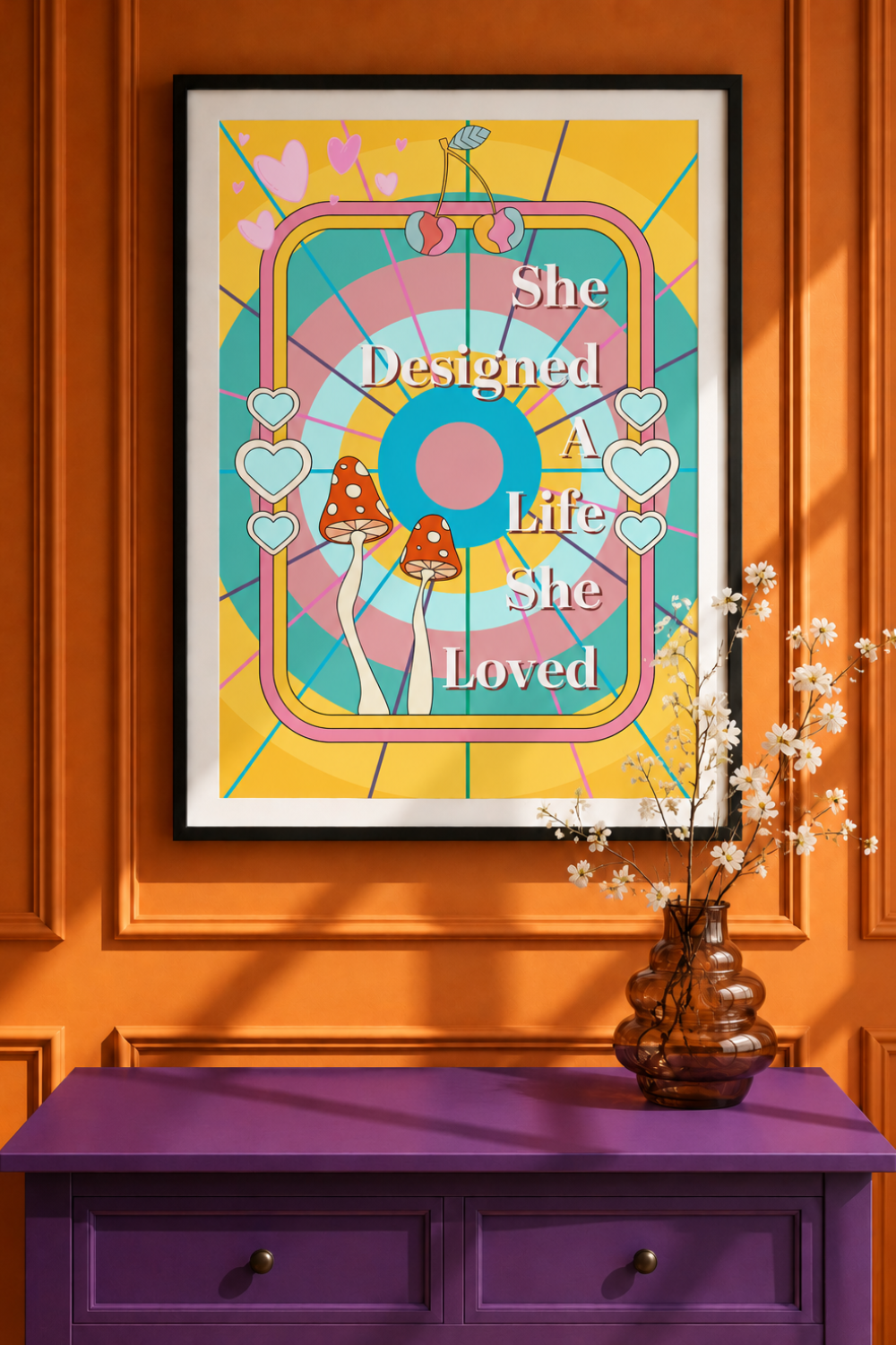 A framed art print featuring a colorful retro design with the text 'She Designed A Life She Loved' and illustrated mushrooms, displayed on an orange wall above a purple cabinet with a vase of flowers.