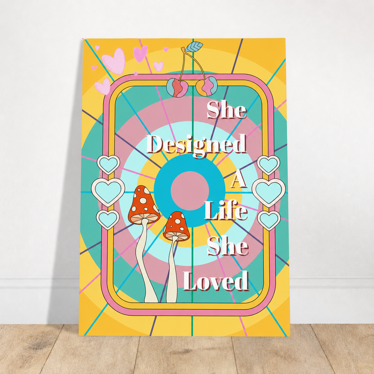Unframed She Designed A Life She Loved art print with colourful circles, hearts, cherries and orange mushrooms, leaning against a white wall on a wooden floor.