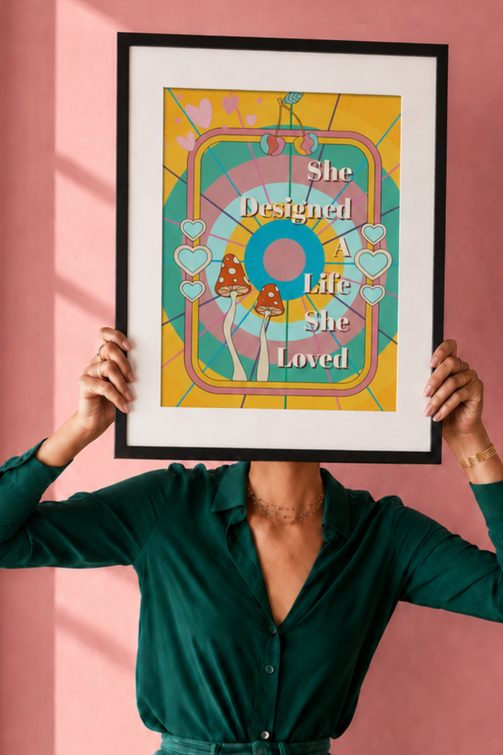 A person in a dark green blouse holds a framed art print in front of their face. The print features a colorful retro design with the text 'She Designed A Life She Loved' over a circular pattern with hearts and mushrooms, set against a pink wall.